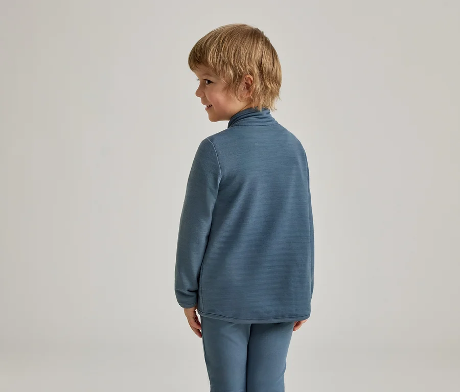 Ein kleines Kind mit blondem Haar, von hinten und der Seite gesehen, trägt ein blaues Langarmshirt mit hohem Kragen und eine passende blaue Hose. Das Kind blickt mit einem leichten Lächeln nach rechts.