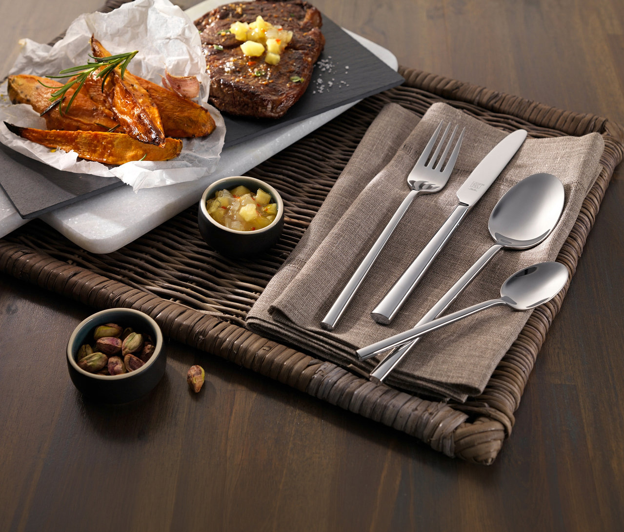 Ein Gericht mit Steak, Süßkartoffelspalten und einem ZWILLING »Aberdeen« Besteck-Set auf einem geflochtenen Tablett. Dazu kleine Schalen mit Salsa und Pistazien.