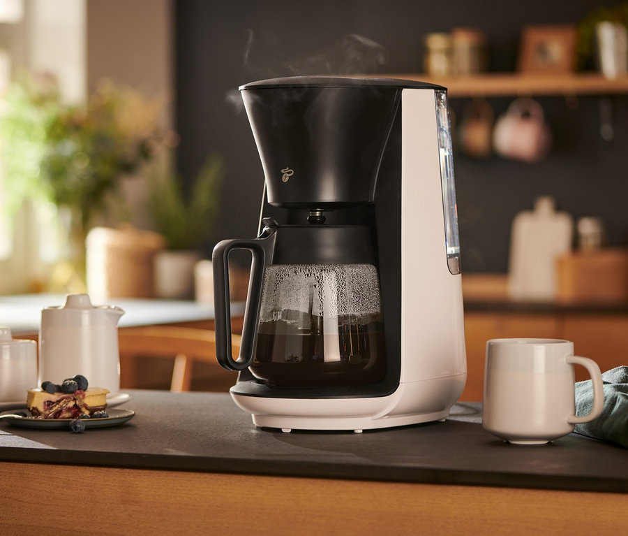 Eine weiße »Let's Brew« Filterkaffeemaschine steht auf einem Tisch neben einer Tasse, einem Milchkännchen, einer Zuckerdose und einem Teller mit Kuchen.