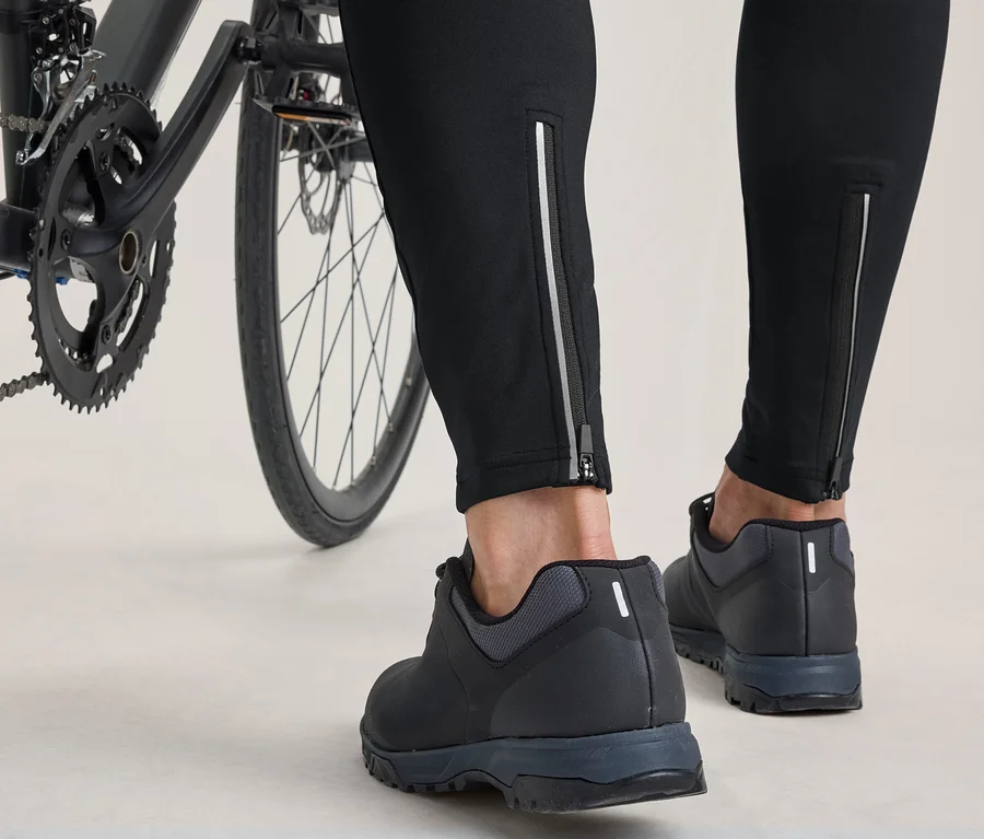 Unterschenkel und Füße einer Person, die neben einem Fahrrad steht. Sie trägt schwarze Hosen mit reflektierenden Reißverschlüssen an den Knöcheln und schwarze Radschuhe.