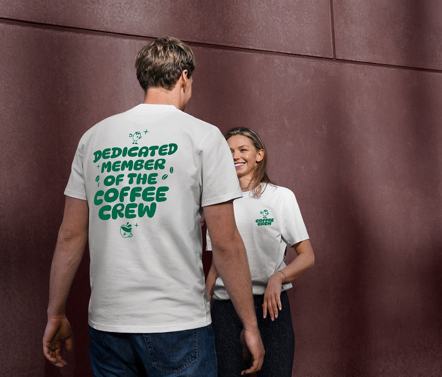 Ein Mann trägt ein weißes Unisex Coffee Crew T-Shirt mit grünem Aufdruck "DEDICATED MEMBER OF THE COFFEE CREW" auf dem Rücken. Eine Frau lächelt im Hintergrund und trägt ebenfalls ein weißes T-Shirt mit dem "Coffee Crew"-Logo.
