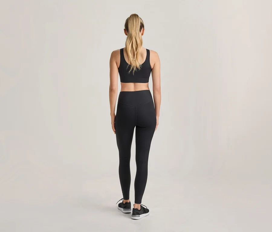 Frau von hinten gesehen, trägt schwarzen Sport-BH, schwarze Leggings und schwarze Sportschuhe, mit blondem Pferdeschwanz.