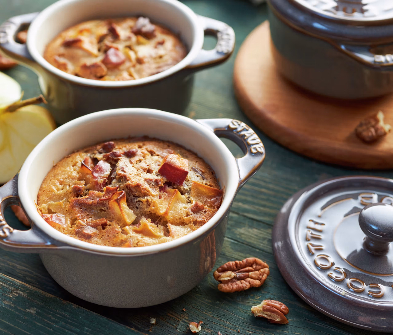 Zwei antikgraue STAUB Keramik-Cocottes mit gebackenem Apfeldessert auf grünem Holztisch, daneben Pekannüsse und ein Apfel.
