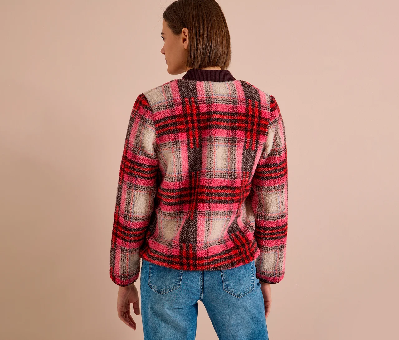Eine Frau von hinten, leicht gedreht, trägt einen karierten CECIL Blouson in Rosa, Rot, Schwarz und Beige aus Fleece-Stoff und blaue Jeans.