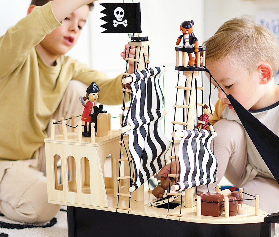 Zwei Jungen spielen mit einem „small foot Piratenschiff“ aus Holz auf einem Teppich.