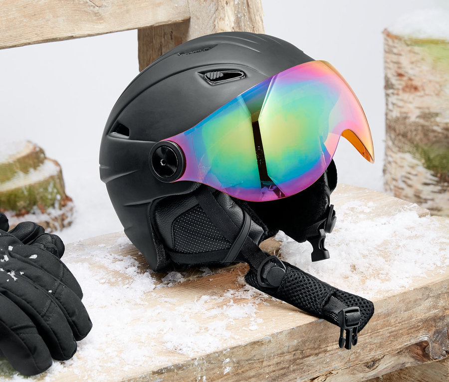 Ein Hardshell-Ski- und Snowboardhelm mit Visier liegt auf einer Holzbank. Daneben liegen schwarze Handschuhe.