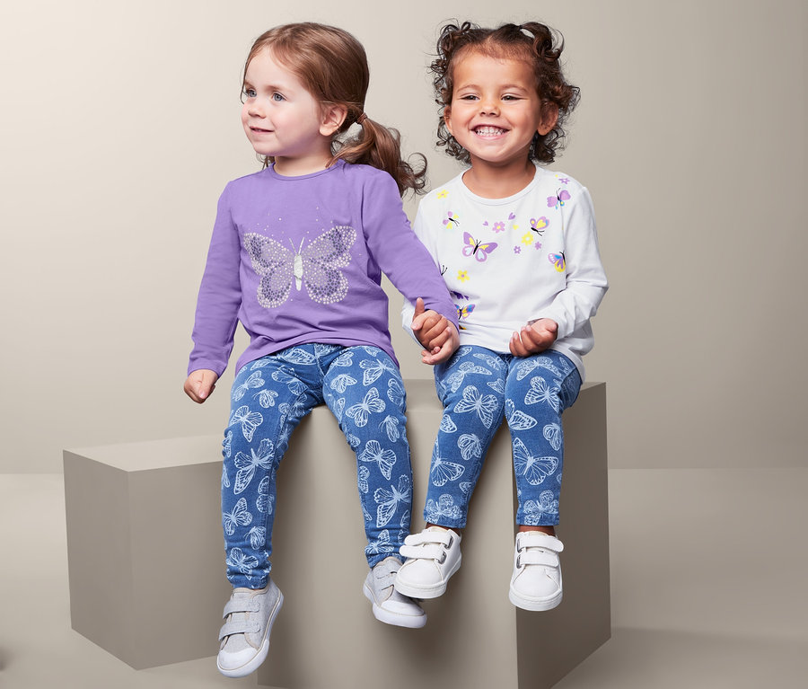 Zwei kleine Mädchen sitzen nebeneinander und halten Händchen. Sie tragen Jeansleggings mit Schmetterlingen und 2 Langarmshirts.