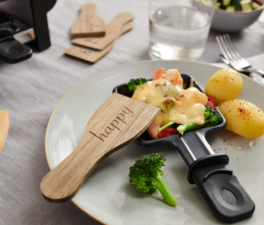 Nahaufnahme einer Raclette-Pfanne mit geschmolzenem Käse, Gemüse und Kartoffeln, mit einem hölzernen 4 Raclette-Schieber mit der Aufschrift "happy".