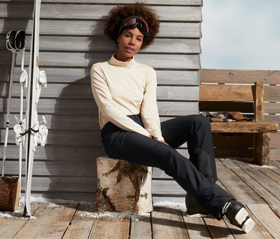 Frau sitzt auf Holzscheit in schwarzer Jetskihose und Skischuhen.