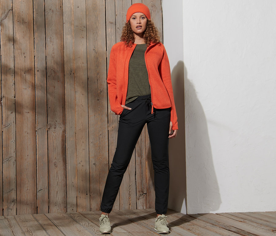 Frau posiert in orangefarbener Jacke und schwarzen Softshell-Joggpants.