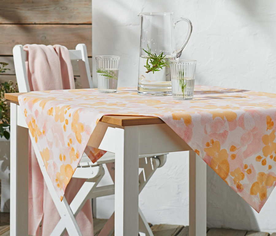 Auf einem weißen Gartentisch liegt eine Outdoor-Mitteldecke, ca. 90 x 90 cm mit orangefarbenem Blumenmuster. Auf dem Tisch stehen ein Glaskrug und zwei Gläser mit Wasser.