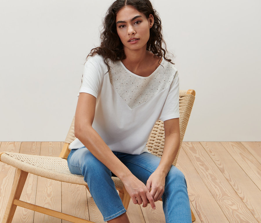 Frau sitzt auf einem Korbsessel und trägt ein weißes Blusenshirt mit Stickerei.
