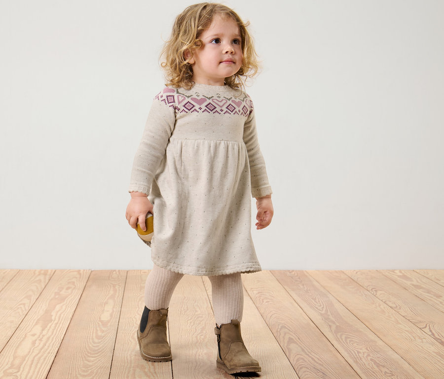 Ein Mädchen steht auf einem Holzboden und trägt ein Kinder-Strick-Kleid.