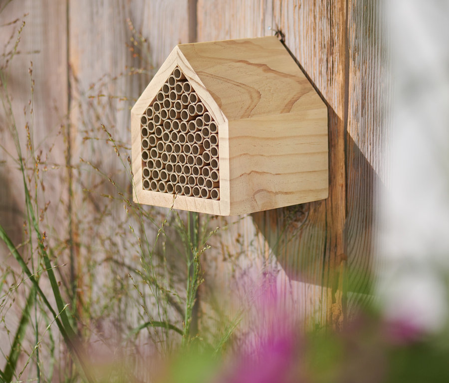 Ein hölzernes Insektenhotel in Hausform ist an einem Holzzaun befestigt.