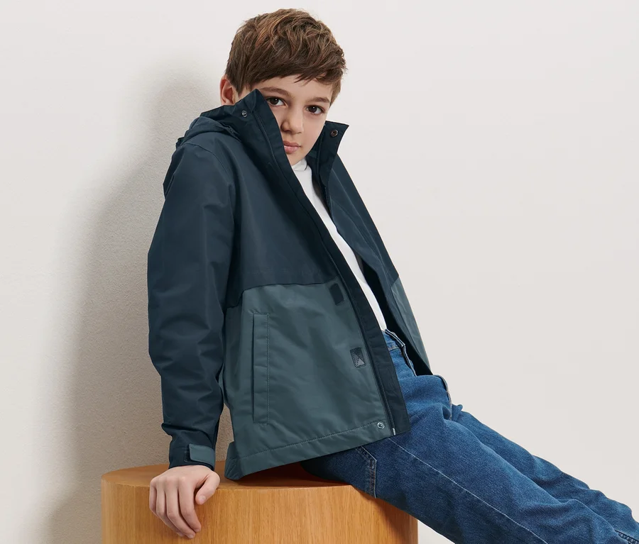 Junge sitzt auf einem Holzhocker und trägt eine Kinder-Allwetterjacke.
