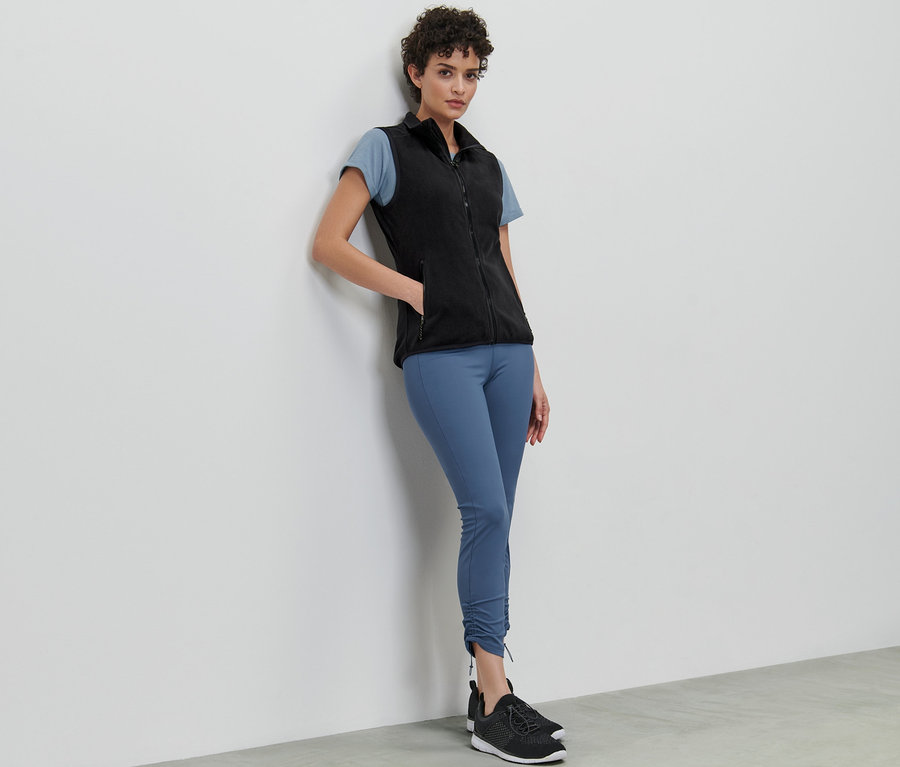 Eine Frau lehnt an einer Wand und trägt eine Fleece-Weste, ein T-Shirt und Leggings.