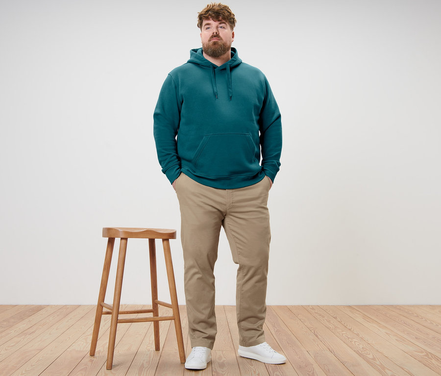 Mann steht mit Händen in den Hosentaschen. Er trägt einen petrolfarbenen Hoodie und eine beige Chino.