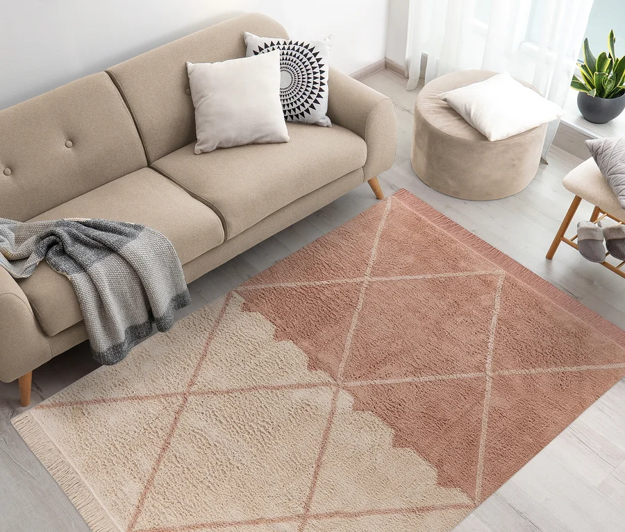 Beiges Sofa mit Kissen und Überwurf, daneben ein beiger Hocker, im Vordergrund der rosa-cremefarbene Teppich Kayoom Mylen.