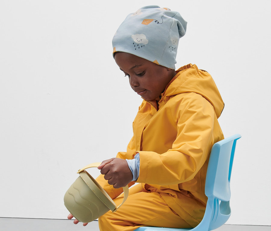 Kind sitzt auf blauem Stuhl, trägt gelben Regenmantel und hellblaue Kinder-Jerseymütze und gießt Wasser aus einem Spielzeugeimer.