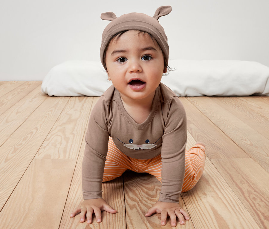 Baby-Set: Baby krabbelt auf Holzboden in braunem Body mit Aufdruck und orange gestreifter Hose. Braune Mütze mit Ohren.