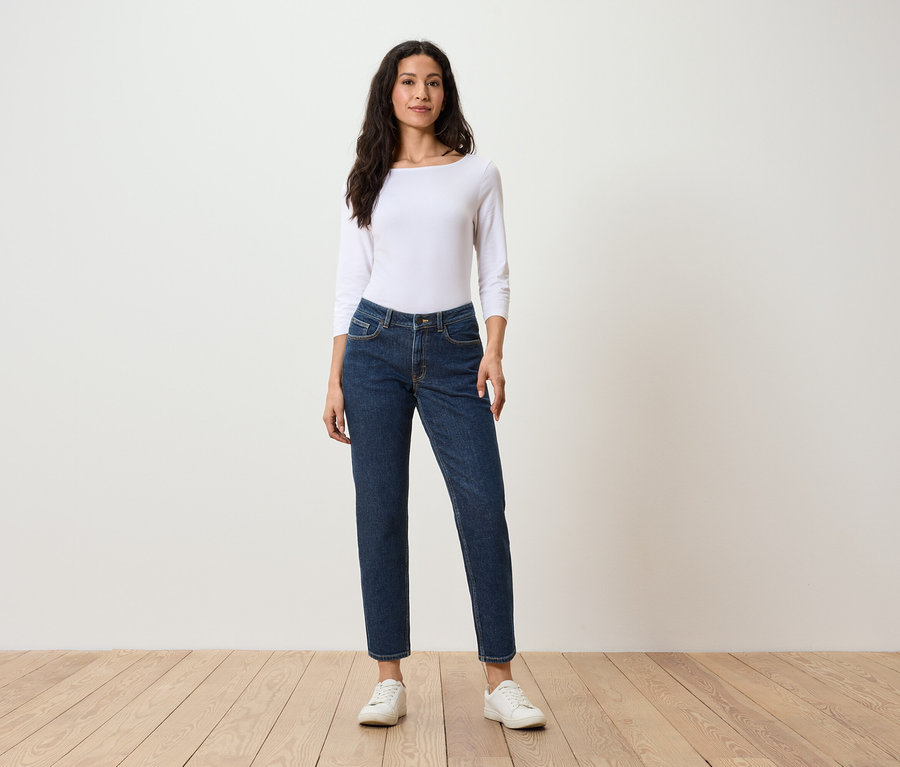 Eine Frau steht und trägt ein weißes Langarmshirt, dunkelblaue Jeans und weiße Turnschuhe.
