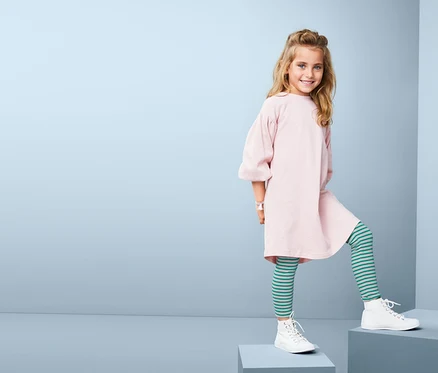 Lächelndes Mädchen posiert auf Stufen in rosa Kleid und grün gestreiften 2 Leggings.