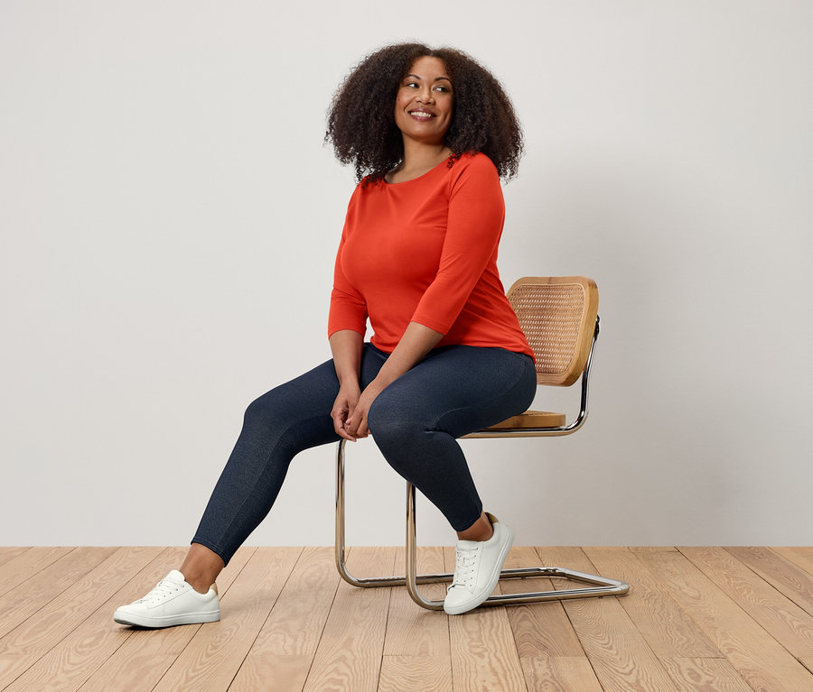 Frau sitzt auf einem Stuhl, trägt eine orangefarbene Bluse, dunkelblaue Leggings und weiße Turnschuhe.