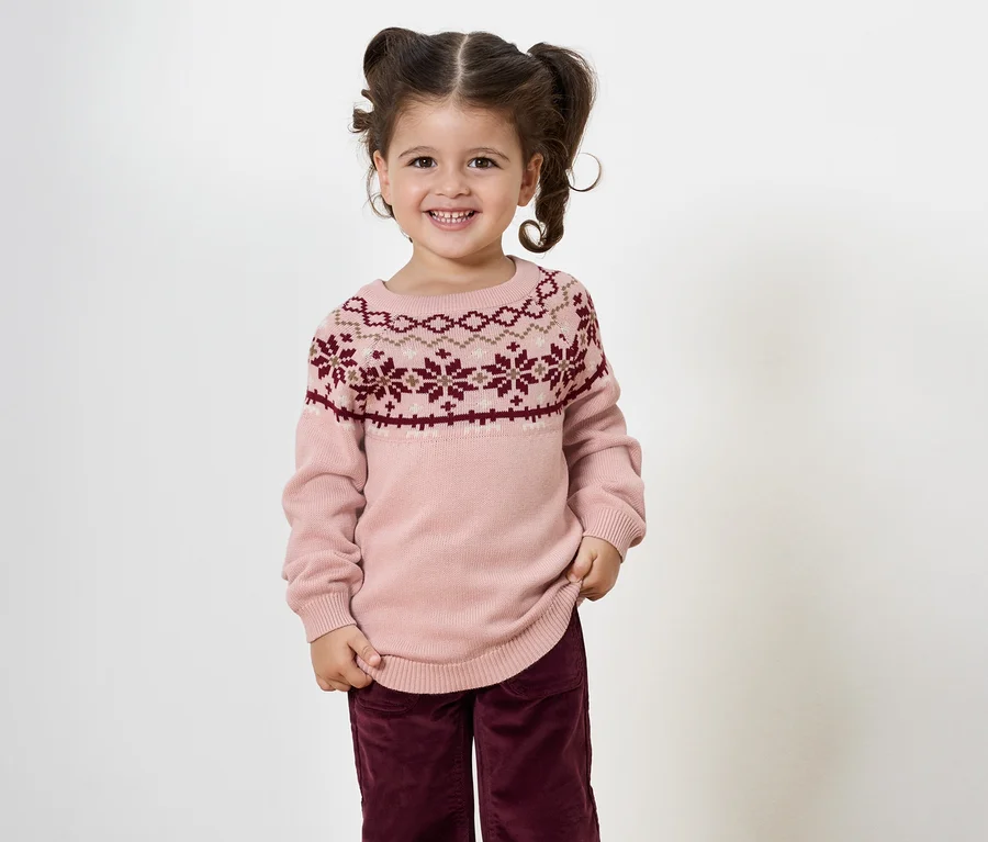 Lächelndes kleines Mädchen mit Zöpfen posiert in einem rosa Pullover mit Muster und weinroten Hosen.