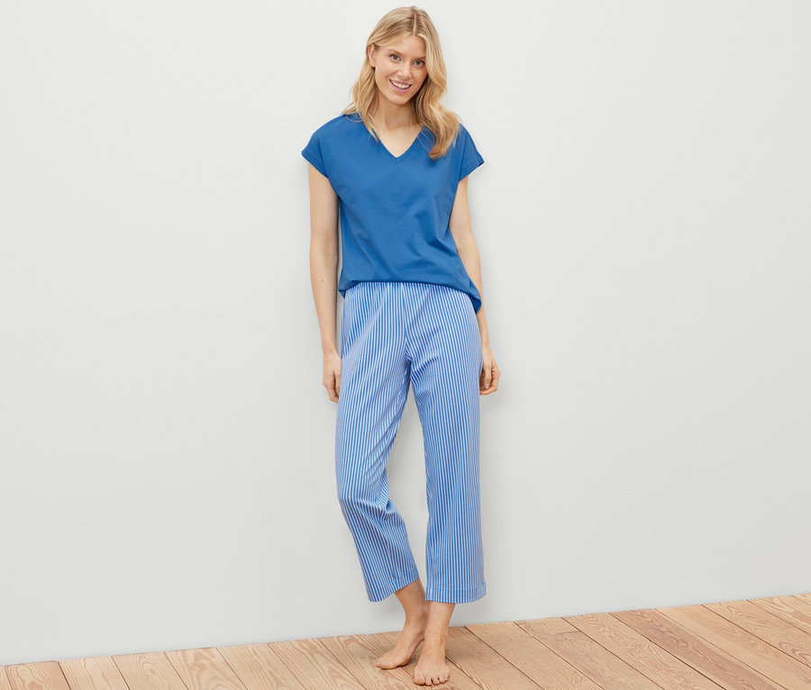 Blonde Frau steht und trägt ein Pyjama-Set, bestehend aus einem blauen T-Shirt und blau-weiß gestreiften Hosen.