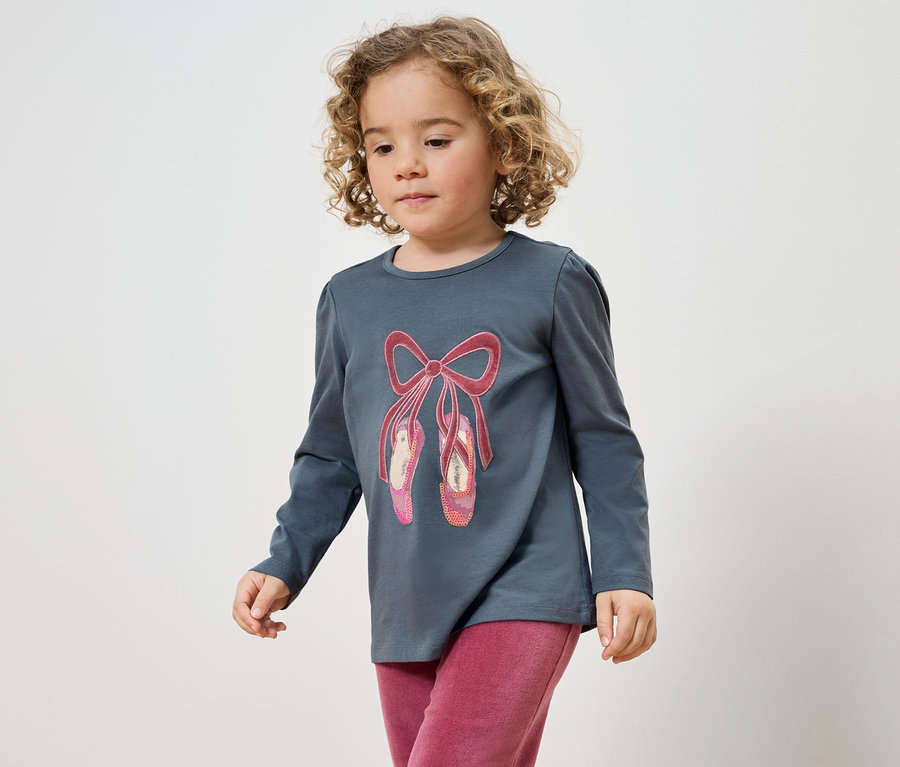 Mädchen mit lockigem Haar trägt ein graues T-Shirt mit einem Ballett-Schuh-Aufdruck und eine pinke Hose.