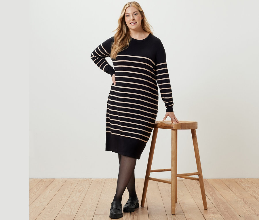 Eine Frau posiert in einem gestreiften Feinstrickkleid neben einem Holzhocker.