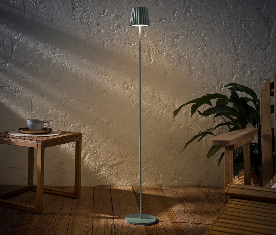 Eine salbeifarbene LED-Akku-Outdoor-Stehleuchte steht neben einem Holztisch mit einer Tasse und einem Holzstuhl.