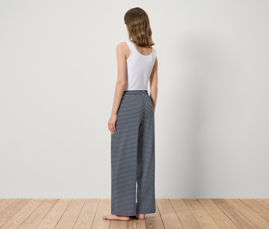 Frau steht mit dem Rücken zur Kamera, trägt ein weißes Top und blau-gestreifte Hosen.