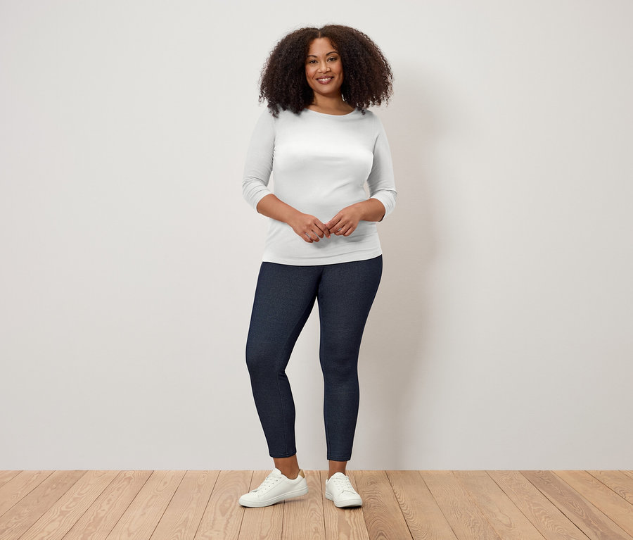 Frau mit lockigem Haar steht auf einem Holzboden und trägt ein weißes Dreiviertelarm-Shirt und dunkelblaue Leggings.