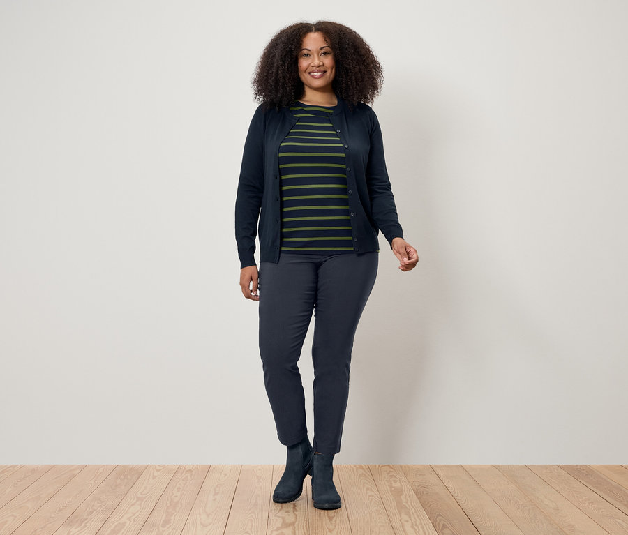 Eine Frau mit lockigem Haar steht auf einem Holzboden vor einer weißen Wand. Sie trägt eine dunkelblaue Strickjacke, ein gestreiftes T-Shirt, dunkelblaue Hosen und Stiefel.