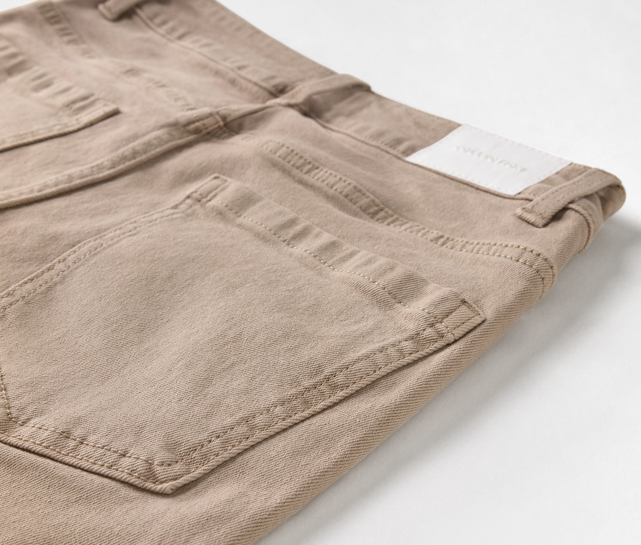 Detailaufnahme einer beigen Jeans mit Taschen und Logo-Patch.