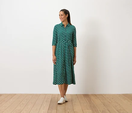 Eine Frau steht und trägt ein grünes gemustertes Kleid und weiße Turnschuhe.