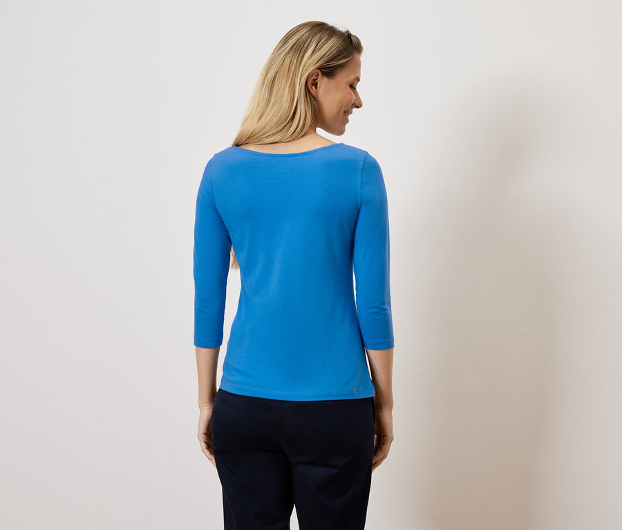 Frau blickt über die Schulter, trägt ein blaues Oberteil mit Dreiviertelärmeln und eine dunkle Hose.