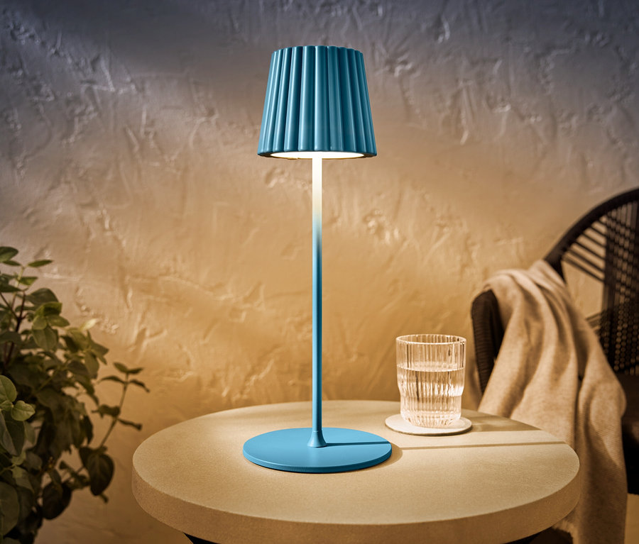 Eine blaue LED-Akku-Outdoor-Tischleuchte steht auf einem Tisch neben einem Glas Wasser.