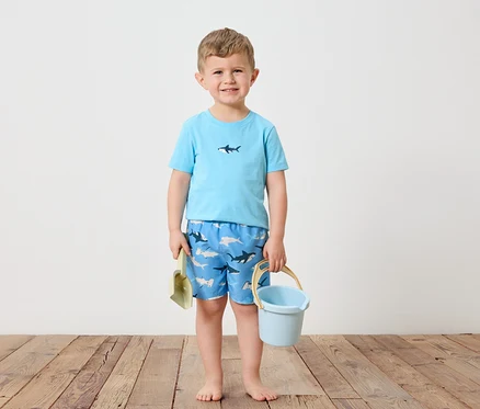 Ein kleiner Junge mit blonden Haaren steht barfuß auf einem Holzboden. Er trägt ein hellblaues T-Shirt mit einem Hai darauf und blaue Badeshorts mit Hai-Motiv. In seiner linken Hand hält er eine grüne Schaufel und in seiner rechten einen hellblauen Eimer.