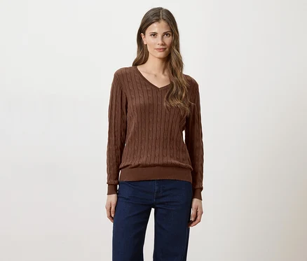 Frau mit langen braunen Haaren, die einen braunen V-Ausschnitt-Pullover und Jeans vor einem weißen Hintergrund trägt.