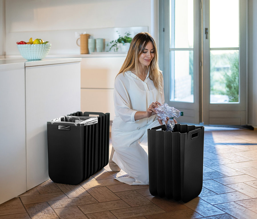 Frau kniet und wirft Plastikflasche in einen von zwei schwarzen Mülleimern im Zimmer.