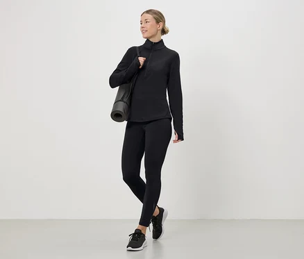 Frau in schwarzem Sportoberteil, Leggings und Turnschuhen mit Yogamatte über der Schulter.