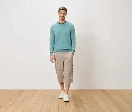 Ein Mann steht mit einer Hand in der Tasche vor einer weißen Wand. Er trägt einen hellblauen Pullover, beigefarbene Dreiviertel-Cargo-Hosen und weiße Schuhe.