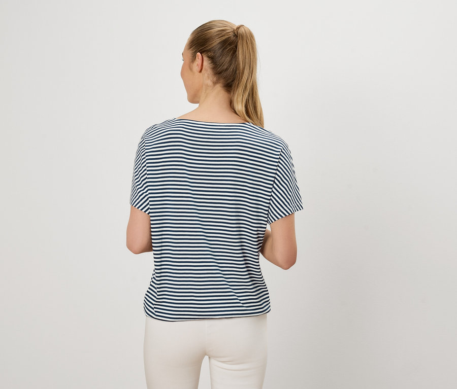 Frau mit blondem Pferdeschwanz trägt ein blau-weiß gestreiftes T-Shirt und helle Hosen, von hinten gesehen.