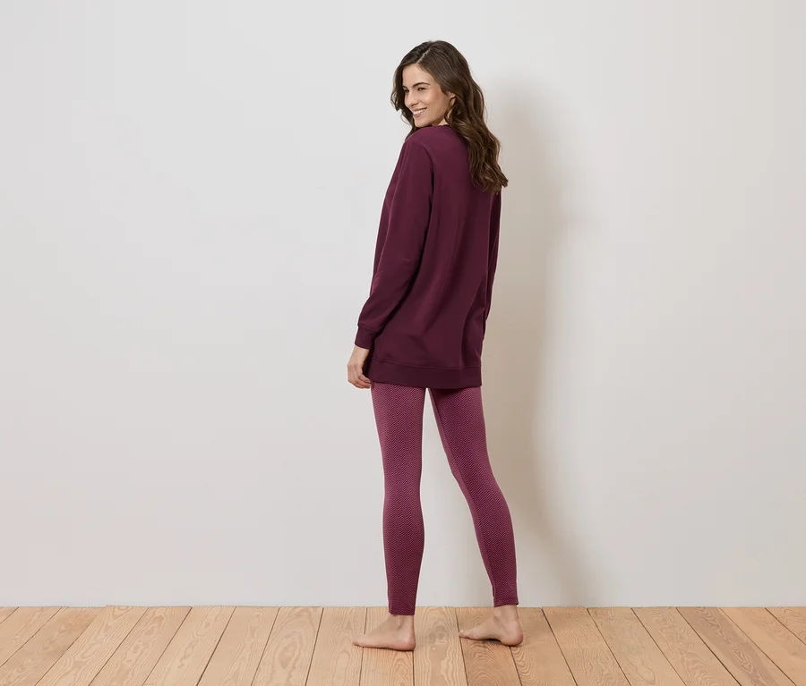 Frau steht barfuß auf einem Holzboden, trägt eine lange, bordeauxrote Bluse und Leggings mit einem geometrischen Muster.