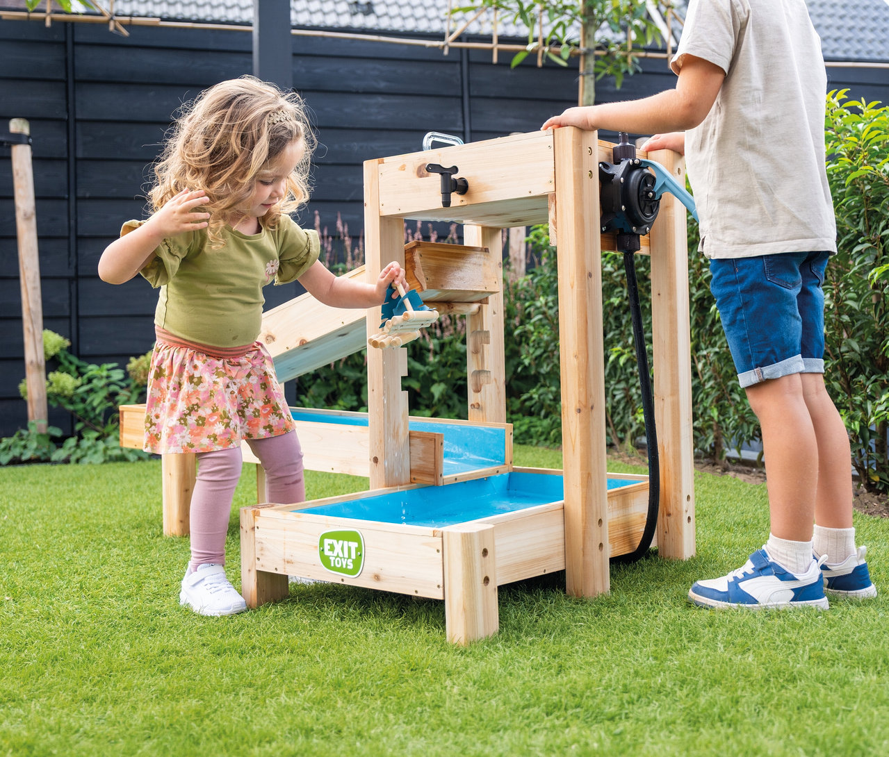 Auf Rasen spielen zwei Kinder mit einem EXIT Toys Wasserspieltisch aus Holz mit blauen Wannen und einer Pumpe.