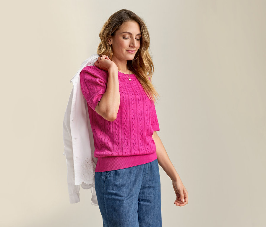 Frau mit Jeansjacke mit Lochstickerei über der Schulter und Feinstrickpullover mit Zopfmuster in Pink zu Jeanshose.