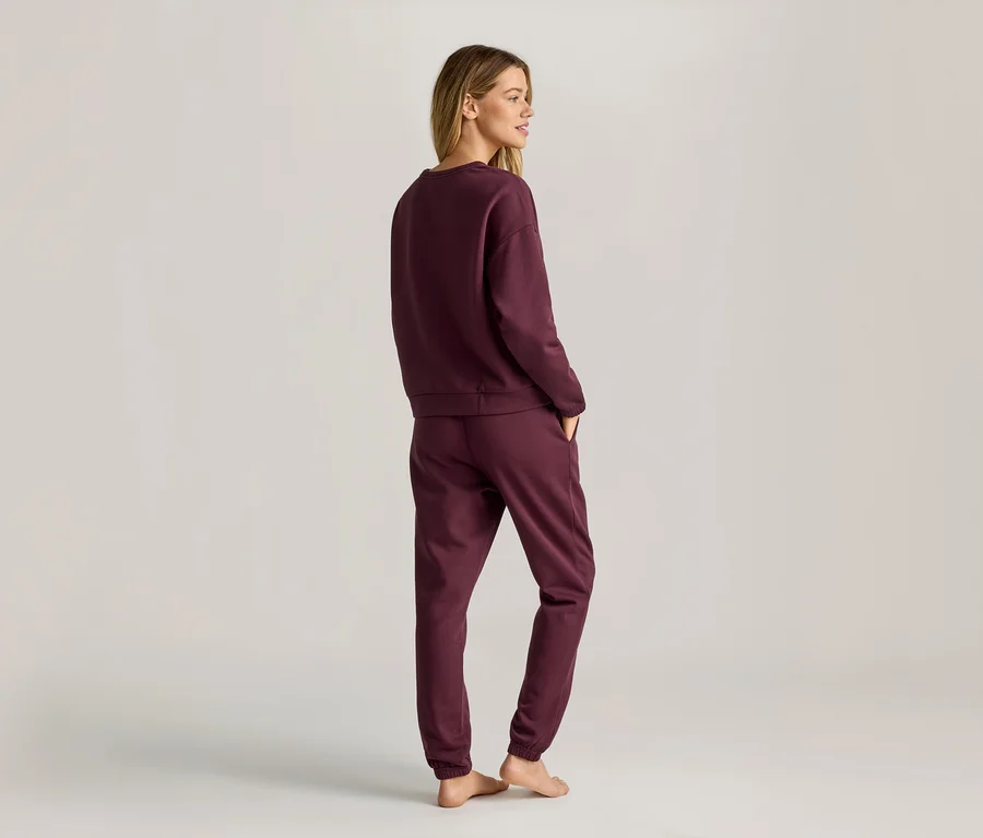 Eine Frau mit blonden Haaren trägt ein weinrotes Sweatshirt und eine Jogginghose, die sie von hinten zeigt.