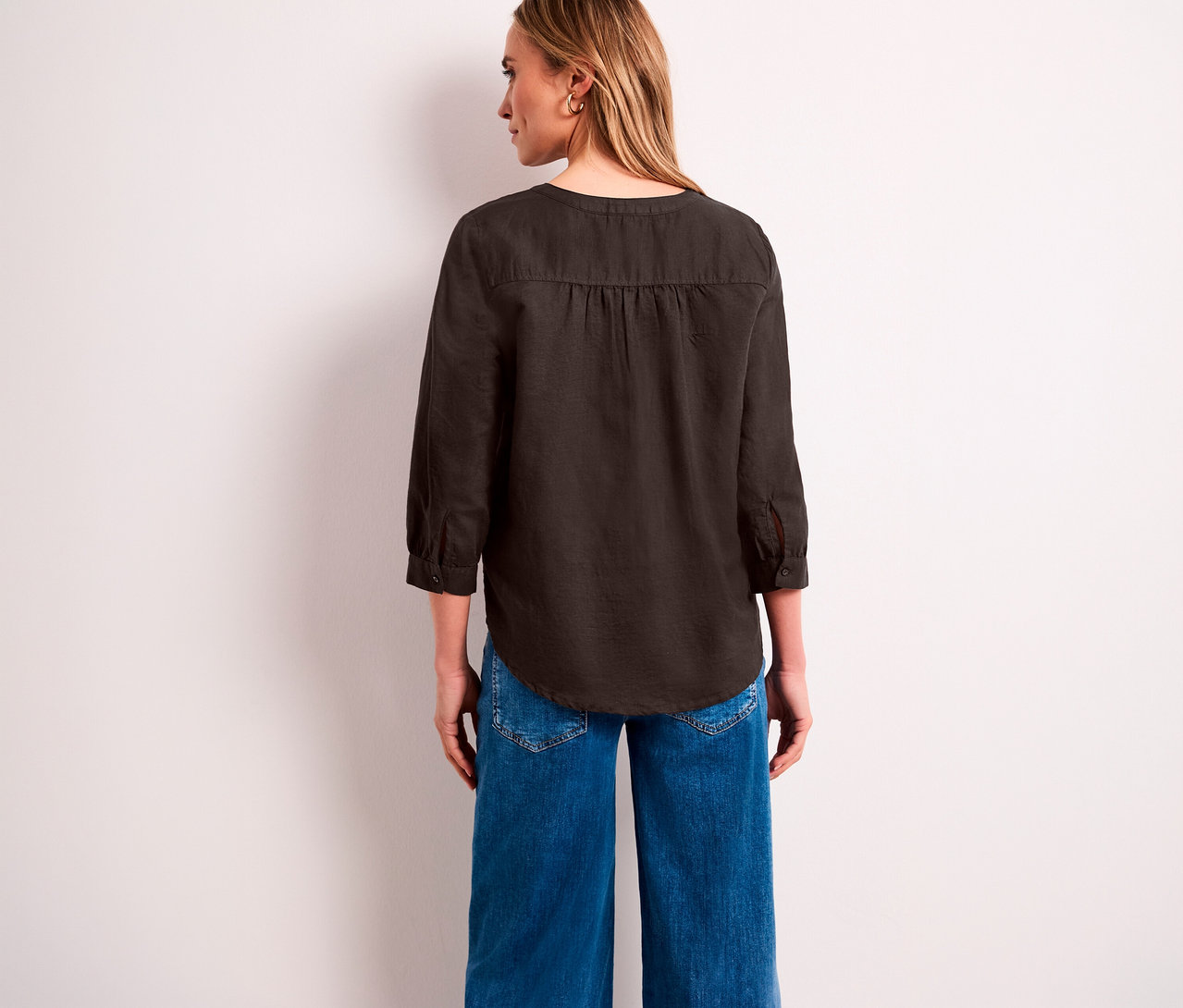 Eine Frau von hinten, nach rechts blickend, trägt eine dunkelbraune Street One Tunika-Bluse mit 3/4-Ärmeln und blaue weite Jeans.